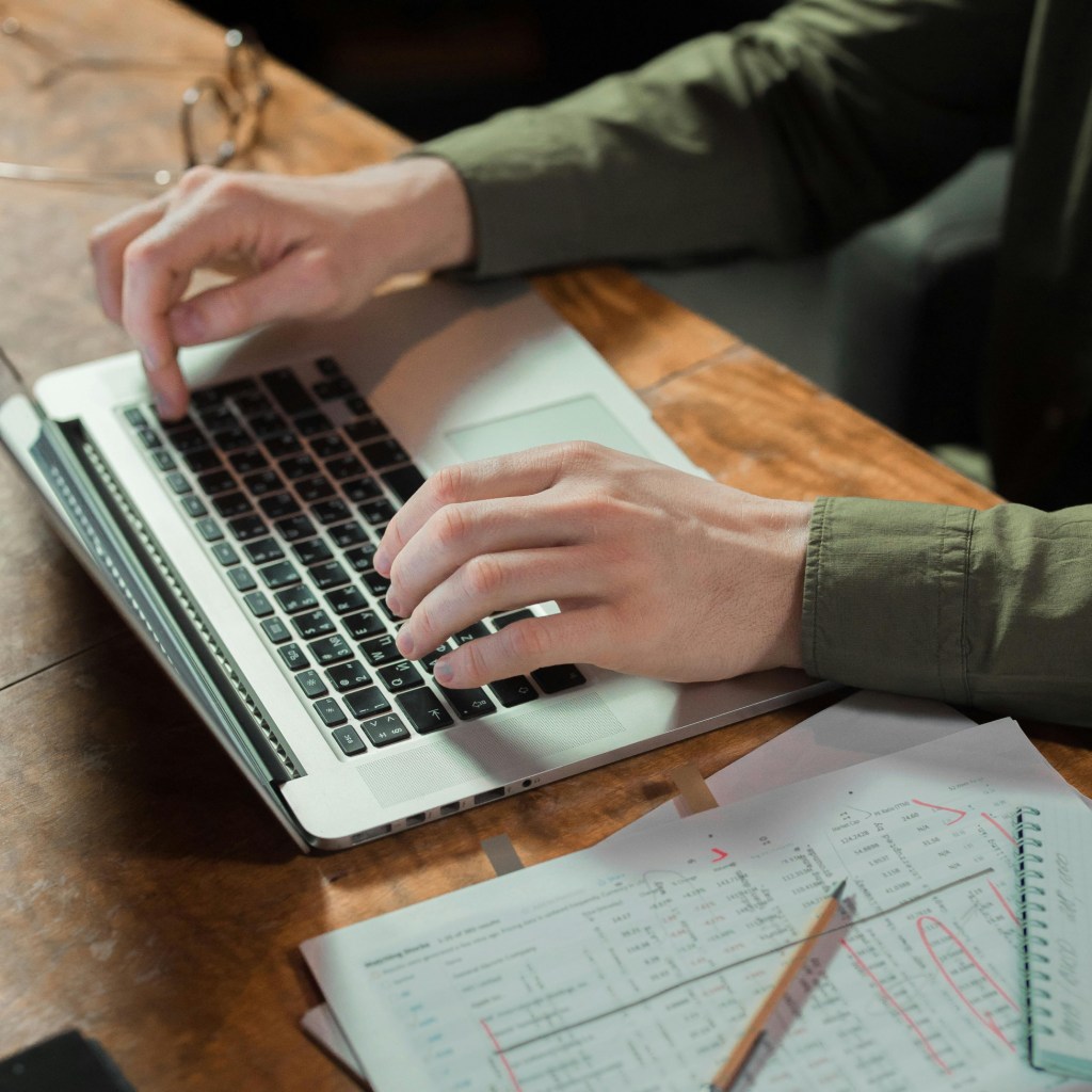 Hands typing on a laptop next to a printed spreadsheet